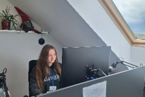 Kobieta siedzi przy stanowisku komputerowym z dużym monitorem i sprzętem elektronicznym. Biurko znajduje się pod skośnym dachem z dużym oknem dachowym, a na ścianie obok stoją rośliny i drobne przedmioty.