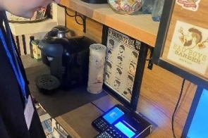 Młody mężczyzna stojący przy laptopie oraz dwóch urządzeniach elektronicznych. Przed nim półka, na której znajduje się ekspres do kawy, butelki wody oraz słoiki.