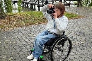 Młoda kobieta na wózku inwalidzkim z plakietką na szyi trzymająca przy oczach aparat fotograficzny. Wózek stoi na brukowanym chodniku, w tle pomost nad jeziorem i wierzby płaczące.