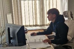 Młody mężczyzna z plakietką siedzący przed komputerem trzymając myszkę. W drugiej ręce trzyma segregator z wpiętymi dokumentami. Obok monitora organizery na dokumenty z przegródkami.
