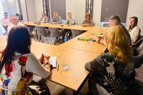 Osoby siedzą przy stołach ustawionych w kształcie podkowy. Na stołach znajdują się dokumenty, notatki i szklanki. Uczestnicy zajmują miejsca wokół stołów i są zwróceni w stronę środka sali