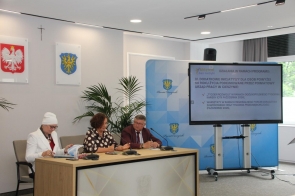 Trzy osoby siedzą przy stole prezydialnym w sali sesyjnej, obok dużego ekranu z prezentacją programu. W tle znajdują się herby, krzyż, rośliny oraz niebieska ścianka z herbem Powiatu Cieszyńskiego.