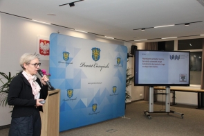 Maria Deskur prowadzi prezentację i mówi do mikrofonu. Obok niej znajduje się plansza Powiatu Cieszyńskiego, a na ekranie wyświetlony jest slajd prezentacji.