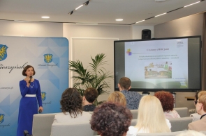 dr Wanda Matras-Mastalerz w niebieskiej sukience prowadzi prezentację przed publicznością. Trzyma mikrofon i stoi obok ekranu, na którym wyświetlony jest slajd.