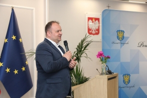 Marcin Szarek Radny Sejmiku Województwa Śląskiego stoi przy mównicy i mówi do mikrofonu. W tle widoczne są flaga Unii Europejskiej, godło Polski, plansza Powiatu Cieszyńskiego oraz rośliny doniczkowe.
