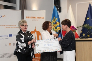 Trzy kobiety podczas uroczystości w sali sesyjnej. Od lewej stoi Beata Macura Członkini Zarządu,obok Janina Żagan Starosta Cieszyński trzyma mikrofon przy Izabeli Kuli Dyrektor Biblioteki Miejskiej w Cieszynie . Kobiety prezentują symboliczny bon. W tle widać banery Biblioteki oraz flagi.