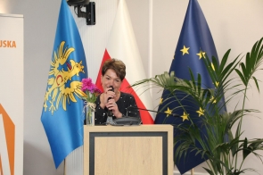 Izabela Kula Dyrektor Biblioteki Miejskiej w Cieszynie  stoi przy mównicy i mówi do mikrofonu. Za nią znajdują się flagi oraz duża roślina doniczkowa. Na mównicy stoi wazon z różowymi kwiatami.