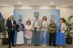 Grupa siedmiu osób pozuje do zdjęcia na tle planszy Powiatu Cieszyńskiego. Część osób trzyma pamiątkowe medale lub etui, a jedna osoba trzyma teczkę. Po bokach widoczne są rośliny doniczkowe.