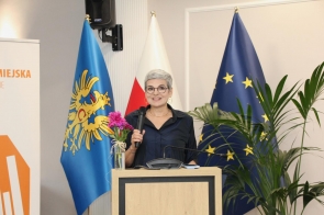 Ewa Katrušák stoi przy mównicy i trzyma mikrofon. W tle widoczne są flagi, rośliny doniczkowe oraz wazon z różowymi kwiatami na mównicy.