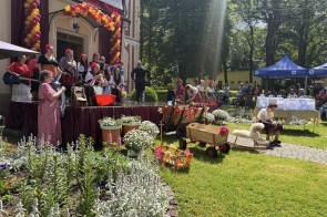 Występ artystyczny w ogrodzie przed budynkiem. Na scenie stoją osoby w kolorowych strojach, a wokół widać dekoracje z balonów, kwiaty, rekwizyty oraz publiczność siedzącą pod namiotem.