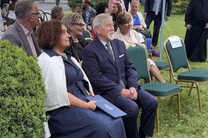 Uczestnicy uroczystości siedzą pod namiotem w ogrodzie. Na pierwszym planie kobieta trzyma teczkę, obok siedzą kolejne osoby, a w tle widać pozostałych gości oraz zieleń.