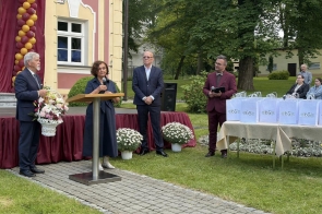 Uroczystość plenerowa przed budynkiem. Kobieta przemawia przy mównicy, obok stoją trzy osoby, a na stole widoczne są torby upominkowe z oznaczeniem 65-lecia.