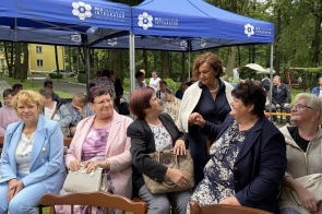 Uczestniczki uroczystości siedzą na ławkach pod niebieskimi namiotami w ogrodzie. Kilka kobiet rozmawia ze sobą, a w tle widać pozostałych gości oraz drzewa.