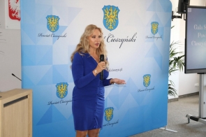 Edyta Pamuła Prezes Wydawnictwa Prasa Beskidzka  w niebieskiej sukience przemawia z mikrofonem na tle baneru z herbem i napisem „Powiat Cieszyński”. Obok widać mównicę oraz ekran z prezentacją gali sportu.