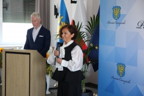 Janina Żagan Starosta Cieszyński przemawia do zgromadzonych, trzymając mikrofon. Obok niej przy mównicy stoi mężczyzna. W tle widać flagi, rośliny oraz baner z herbem.