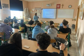 Uczniowie siedzą w ławkach w sali lekcyjnej. Z przodu stoi osoba prowadząca, obok ekranu z wyświetloną prezentacją.