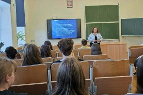 Uczniowie siedzą w sali wykładowej. Z przodu stoi osoba prowadząca, obok ekranu z wyświetloną prezentacją i tablic szkolnych.
