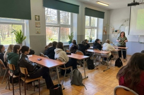 Uczniowie siedzą przy ławkach w sali lekcyjnej. Z przodu stoi osoba prowadząca, obok ekranu z wyświetloną prezentacją.