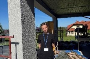 Młoda kobieta stoi na zewnątrz przy budynku w trakcie prac budowlanych. W tle widać rusztowanie, materiały budowlane i fragment zadaszenia.
