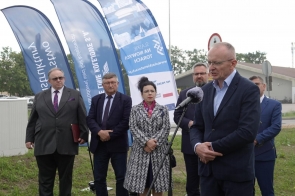 Mężczyzna przemawia do mikrofonu na zewnątrz. Za nim stoją cztery osoby oraz niebieskie flagi promocyjne, a w tle widać ulicę i budynki.