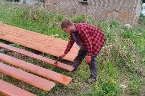 Młody mężczyzna pracuje na zewnątrz przy drewnianych deskach ułożonych na trawie. W tle widać budynek z cegły oraz zarośnięty teren.