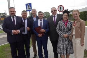 Grupa ośmiu osób pozuje do zdjęcia na chodniku przy drodze. W tle widać znaki drogowe, latarnie, samochody i zachmurzone niebo.