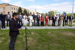 Mężczyzna stoi przy mikrofonie na zewnątrz i trzyma dokumenty. W tle grupa osób stoi na chodniku przy ulicy, obok trawnika i budynków.