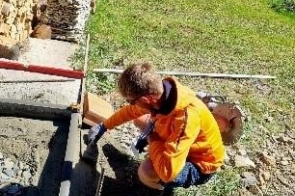 Młody mężczyzna wykonuje prace na zewnątrz przy betonowej wylewce. Ma na sobie odzież roboczą, rękawice i trzyma narzędzie.