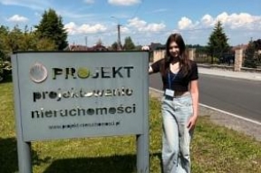 Młoda kobieta stoi obok tablicy z napisem „PROJEKT projektowanie nieruchomości”. W tle widać drogę, trawnik, zabudowania i niebo.