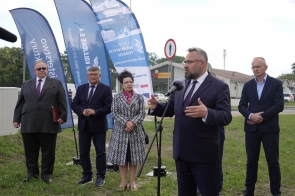 Mężczyzna przemawia do mikrofonu na zewnątrz. Za nim stoją cztery osoby oraz niebieskie flagi promocyjne, a w tle widać ulicę i budynki.