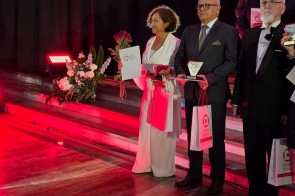 Osoby stoją na scenie z dyplomami, statuetkami, kwiatami i torbami prezentowymi. W tle widać czarną kurtynę, dekoracje kwiatowe oraz czerwone oświetlenie sceniczne.