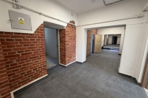 Korytarz po remoncie z szarą posadzką, białymi ścianami i fragmentami odsłoniętej cegły. Widać kilka przejść, drzwi, instalacje pod sufitem oraz oświetlenie.