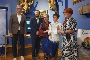 Chłopiec trzyma dyplom i torbę prezentową, pozując do zdjęcia z dorosłymi w eleganckiej sali. W tle widać niebieskie ściany, obrazy i dekoracyjne rzeźby.