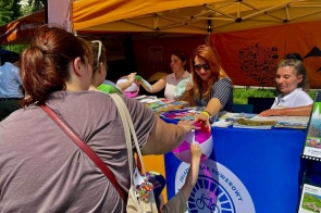 Stoisko informacyjne pod pomarańczowym namiotem, z ulotkami i materiałami promocyjnymi na stole. Przy stoisku stoją osoby, a dziecko trzyma kolorową piłkę plażową.