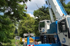 Na drodze widać duży dźwig z wysuniętym ramieniem oraz sprzęt budowlany. Teren prac jest częściowo odgrodzony, a wokół znajdują się drzewa, zieleń i znaki drogowe.