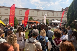 Grupa osób stoi na trawniku podczas plenerowego wydarzenia. Przed nimi widać osoby w historycznych strojach z tarczami, a w tle czerwone flagi, food truck, namiot oraz zabytkowy budynek za ogrodzeniem.
