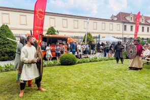 Osoby w historycznych strojach stoją na trawniku podczas plenerowego wydarzenia. W tle widać uczestników, czerwone flagi, food truck, namiot oraz zabytkowy budynek za ogrodzeniem.