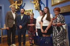 Grupa osób pozuje do zdjęcia w eleganckiej sali z niebieskimi ścianami. Dwie dziewczynki stoją z dyplomem i dużą torbą prezentową, a w tle widać obrazy oraz złote rzeźby.