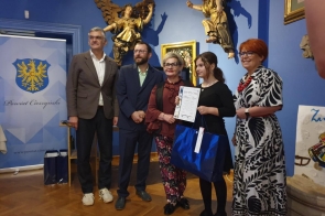 Grupa osób pozuje do zdjęcia w eleganckiej sali z niebieskimi ścianami. Dziewczynka trzyma dyplom i dużą torbę prezentową, a w tle widać baner Powiatu Cieszyńskiego oraz złote rzeźby.