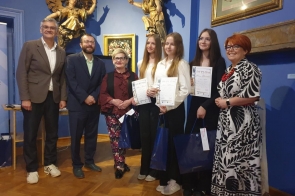 Grupa osób pozuje do zdjęcia w eleganckiej sali z niebieskimi ścianami i złotymi rzeźbami. Trzy dziewczyny trzymają dyplomy i torby prezentowe.