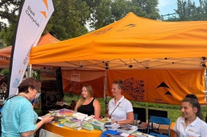 Stoisko informacyjne pod pomarańczowym namiotem z materiałami promocyjnymi i ulotkami. Przy stoisku stoją i siedzą osoby, a obok widoczna jest flaga z napisem „Śląsk Cieszyński”.