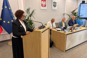 Janina Żagan Starosta Cieszyński stoi przy mównicy i przemawia do mikrofonu w sali obrad. Przy stole prezydialnym siedzą trzy osoby, a w tle widać flagi, godło Polski, herb Powiatu Cieszyńskiego oraz ekran.