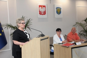Beata Macura Członkini Zarządu stoi przy mównicy i przemawia do mikrofonu. Przy stole obok siedzą dwie osoby, a w tle widać godło Polski, herb Powiatu Cieszyńskiego oraz rośliny.