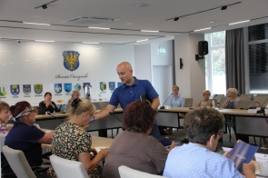 Kamil Waleczek Prawnik rozdaje materiały uczestniczkom siedzącym przy stołach w sali obrad. W tle widać herb i napisy „Powiat Cieszyński” oraz pozostałe osoby siedzące przy stołach.
