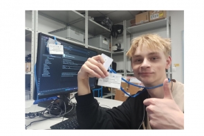 Młoda osoba trzyma niewielki element elektroniczny w pracowni technicznej. W tle widać monitor z kodem oraz wyposażenie stanowiska komputerowego.