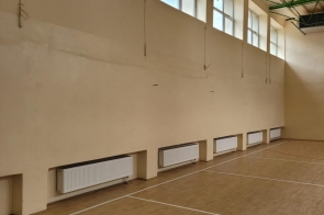 Pusta sala gimnastyczna z drewnianą podłogą i liniami boiska. Przy ścianie widać grzejniki, wysokie okna oraz zieloną konstrukcję dachu.