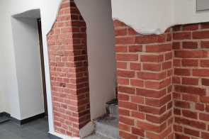 Korytarz z szarą posadzką i białymi ścianami, na których widoczne są fragmenty odsłoniętej czerwonej cegły. W środku znajduje się przejście ze schodami, a pod sufitem widać instalacje.