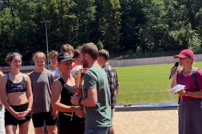 Na stadionie odbywa się wręczenie nagrody. Kobieta otrzymuje puchar, obok stoją uczestnicy biegu, a po prawej stronie osoba prowadząca trzyma mikrofon i kartki. W tle widać boisko, drzewa oraz słoneczne niebo.