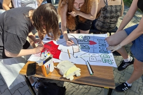 Młodzież wspólnie przygotowuje plakat przy stoliku na zewnątrz. Na arkuszu widoczny jest napis „Bieg po zdrowie” oraz rysunek serca, a obok leżą markery, telefon i pluszowa zabawka.