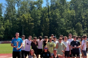 Grupa młodzieży pozuje do wspólnego zdjęcia na bieżni lekkoatletycznej. Część osób trzyma medale i nagrody, a w tle widać drzewa oraz słoneczne niebo.